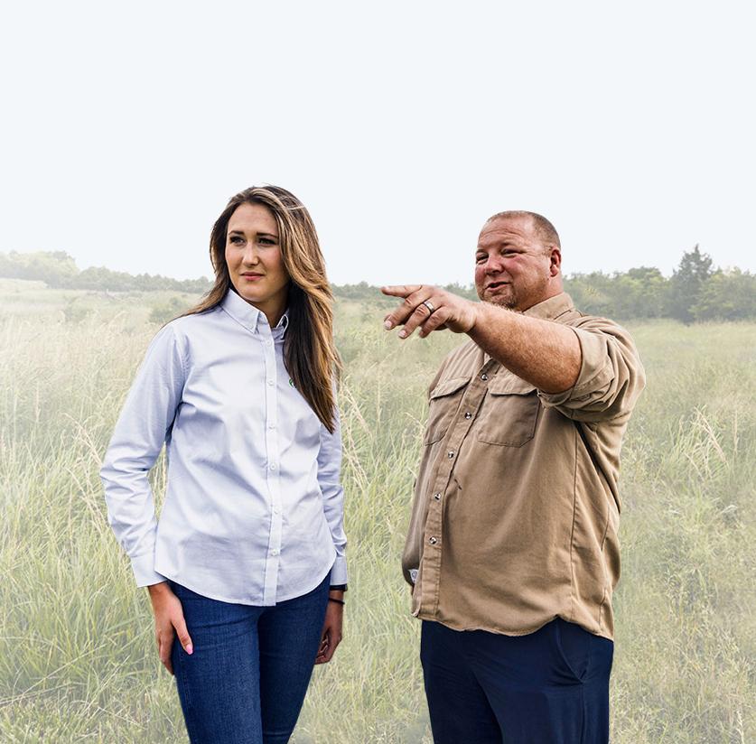 A man points toward the horizon while a woman listens, both standing in a grassy field.